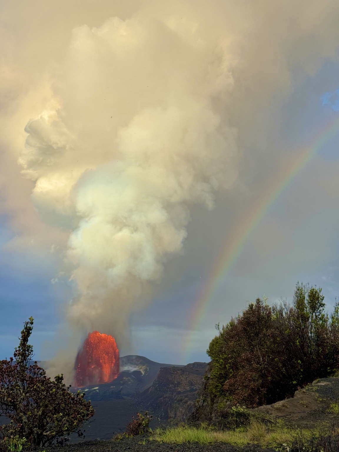 Rafael’s photograph of a rainbow beside a Kīlauea lava fountain and rising eruption plume